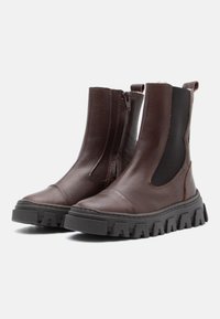 Botas de tobillo de cuero marrón con paneles laterales elásticos, cierre de cremallera y suelas de goma negra gruesa con un patrón dentado.