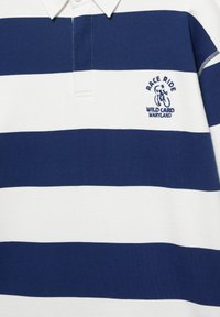 Tengerészkék-fehér, vízszintes csíkos rugby póló fehér gallérral, mellrészen a "Race Ride Wild Card Maryland" kerékpáros logóval hímezve.