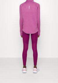 Personne de dos portant un haut à manches longues violet, des leggings magenta ajustés et des baskets blanches avec des accents roses.