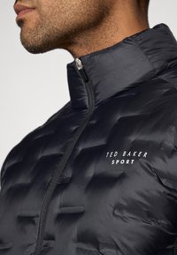Μαύρο μπουφάν πούφ με ψηλό γιακά, με πλισέ υφή, πλήρη φερμουάρ και λογότυπο "TED BAKER SPORT" σε λευκό χρώμα στο στήθος.