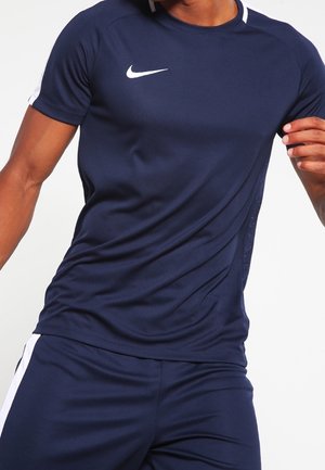 Athlet trägt ein dunkelblaues Nike Sportshirt und passende Shorts, posiert mit leicht angewinkelten Armen vor einem einfarbigen Hintergrund.