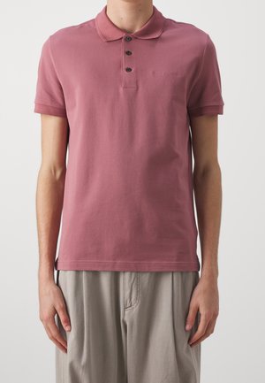 Mann trägt ein kurzärmeliges Polohemd in staubigem Rosé mit drei Knöpfen und hellbeige Hose vor einfachem Hintergrund.