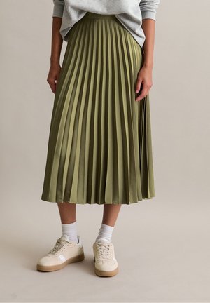 Person iført en olivengrøn plisseret midilang nederdel kombineret med hvide sokker og beige sneakers, stående mod en neutral baggrund.