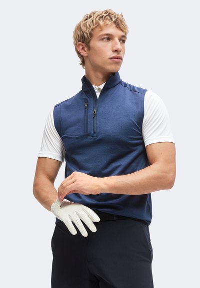 Jeune homme portant un gilet bleu sans manches par-dessus une chemise blanche à manches courtes, enfilant un gant de golf blanc, regardant sur le côté.