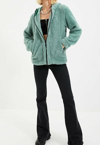 Felpa con zip in pile verde menta, dotata di cappuccio, due tasche frontali e superficie testurizzata. Indossata con pantaloni a zampa neri.