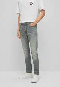 Vaqueros de mezclilla gris claro con un acabado desgastado, cinco bolsillos y trabillas para cinturón, combinados con una camiseta blanca y zapatillas blancas.