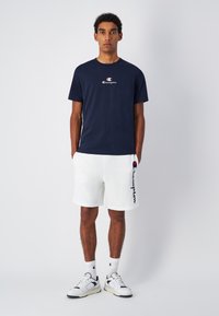 Camiseta de manga corta azul marino con un logo en el pecho, combinada con pantalones cortos blancos que muestran un logo en el lateral. El modelo se encuentra en un fondo neutro.