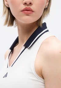 Κοντινό πλάνο ενός ατόμου που φοράει λευκό αμάνικο πουκάμισο με ναυτικό γιακά και λογότυπο τριών μαύρων ρίγων στο στήθος.