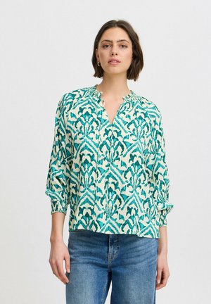 Grün-blau gemusterte Bluse mit einem ausgeschnittenen Ausschnitt und gerafften langen Ärmeln, kombiniert mit blauen Jeans. Glatte Stoffstruktur.