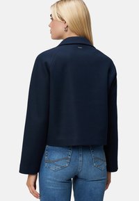 Navy cropped jacket in a textured fabric with a collar, featuring a subtle logo detail and relaxed fit sleeves, paired with fitted jeans.