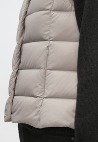 Vestă matlasată de culoare bej deschis, cu design acolorat, având un buzunar cu fermoar pe lateral, purtată peste un pulover negru texturat.