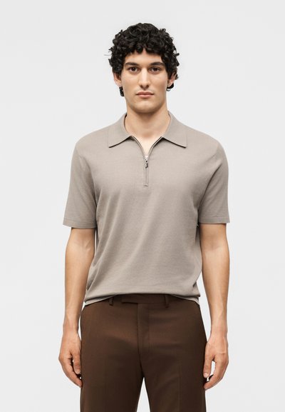 Homme aux cheveux bouclés et foncés portant un polo beige à manches courtes zippé et un pantalon marron, debout devant un fond clair uni.