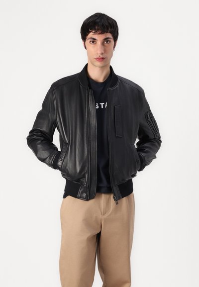 Veste bomber en cuir noir avec fermeture éclair à l'avant, poignets et col côtelés. Présente une poche sur le côté et une texture lisse. Portée sur une chemise bleu marine.