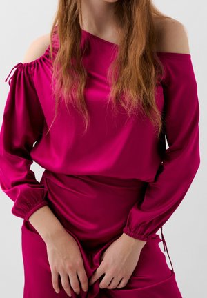 Mujer con cabello largo que lleva una blusa magenta de hombros descubiertos y una falda a juego, con las manos reposando al frente sobre un fondo liso.