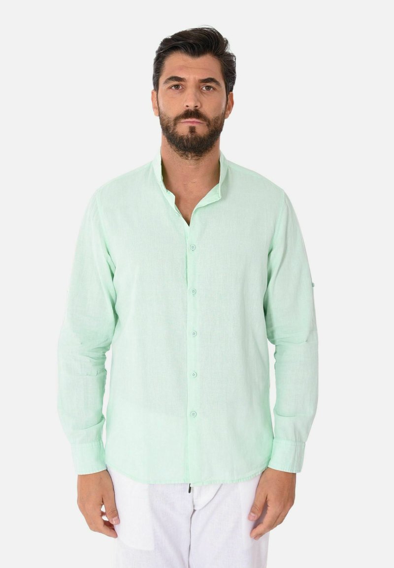 Camicia verde chiaro a maniche lunghe con colletto a bottone e bottoni frontali, realizzata in un tessuto testurizzato; indossata con pantaloni bianchi.