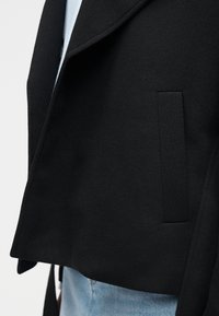 Gros plan sur la poche et l'ourlet d'un manteau en laine noire, par-dessus un jean bleu clair et une chemise bleu clair.