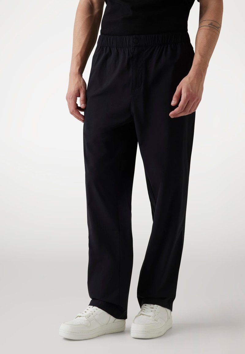 Black elastic waist pants with a relaxed fit, made from lightweight fabric. Features front pockets and is paired with white sneakers.