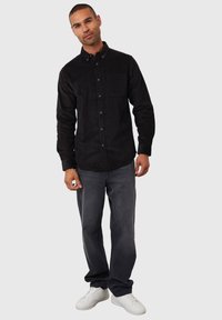 Sort skjorte med knaplukning og en lomme, parret med grå jeans og hvide sneakers. Skjorten har et tekstureret stof og en krave.