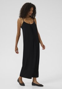 Robe maxi noire avec fines bretelles, décolleté froncé, tissu doux et fentes latérales pour faciliter le mouvement, associée à des chaussures plates noires.