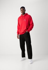 Rød bomulds-hættetrøje med kængurulomme og snoretræk, parret med sorte bukser og hvide sneakers på en neutral baggrund.