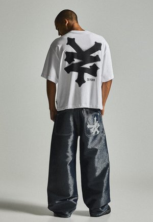 Homme portant un T-shirt blanc oversize avec un logo abstrait noir et un jean large foncé avec un logo assorti sur la poche arrière, debout de dos.