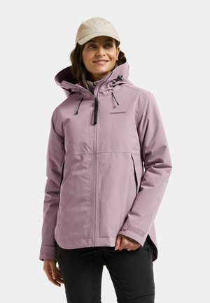 Woman wearing a light purple hooded jacket and beige cap, standing and looking to the side with a slight smile against a plain background.