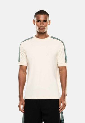 Uomo che indossa una camicia crema a maniche corte con dettagli a strisce verde scuro sulle spalle e pantaloni neri con strisce coordinate.