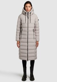 Lys grå lang dunjakke med hætte, der har vertikal quiltning, fuld frontlynlås og sidelommer. Båret over sorte leggings og støvler.
