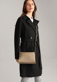 Cappotto trench nero doppiopetto con cintura, abbinato a una borsa a tracolla beige testurizzata con un motivo sottile e tracolla regolabile.