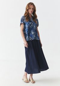 Kurze Ärmel, blaues, tropisches Musterhemd aus leichtem Stoff, kombiniert mit einem langen, marineblauen Leinenrock und beigen Rutschen-Sandalen.