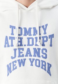 Sudadera blanca con un gran texto azul que dice "TOMMY ATH. DEPT JEANS NEW YORK", con capucha y cordones. Textura de tela suave.
