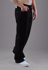 Schwarze Jeans mit geradem Bein, Kupferriegel und Seitentaschen. Kombiniert mit weißen Sneakern, auf einem neutralen Hintergrund präsentiert.