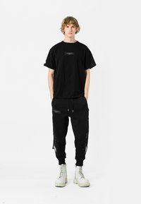 Camiseta de algodón negra con un parche de logo rectangular, combinada con joggers negros que cuentan con cremalleras laterales y detalles de flecos. Botas blancas completan el look.