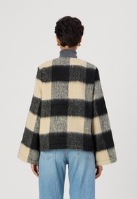 Veste en laine à carreaux noir, crème et gris avec une coupe ample et des manches larges, portée sur un jean bleu clair. Tissu doux et texturé.