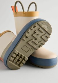 Bottes en caoutchouc avec une tige crème, des accents bleus et beiges, une semelle texturée avec un motif en zigzag et des anses dans des teintes douces.