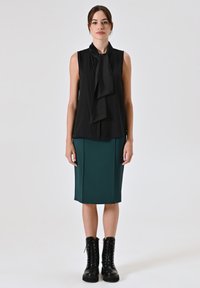 Zwarte mouwloze blouse met een gedrapeerde strikdetail, gecombineerd met een aansluitende groene kokerrok. Draagt zwarte veterschoenen met een chunky zool.