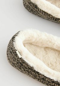 Black and tan herringbone pattern slipper with a soft white faux fur lining, featuring a small label on the side. Round shape and cushioned design.