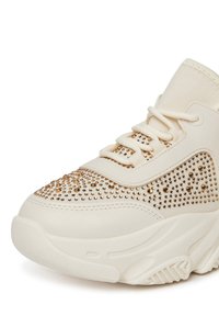 Kremowe trampki na wysokim podeście z grubą podeszwą, zawiązywane z przodu, ozdobione złotymi ćwiekami na palcach i bokach.
