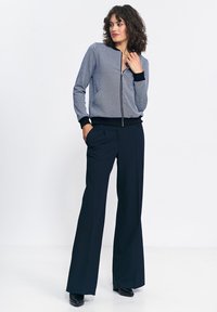 Kobieta z kręconymi włosami, ubrana w szary dres z zamkiem i szerokie granatowe spodnie, stoi z jedną ręką w kieszeni i patrzy na bok.