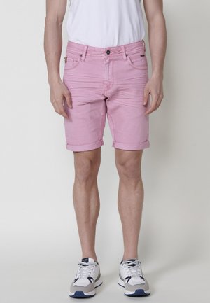 Shorts de mezclilla rosa con dobladillos enrollados, diseño de cinco bolsillos y cierre de cremallera, combinados con una camiseta blanca y zapatillas deportivas.