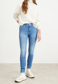 Jeans ajustados de denim en un azul claro, con una cintura de altura media y un ligero desgaste. Combinados con un suéter blanco de punto grueso y zapatillas altas blancas.