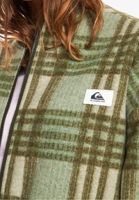 Fleecová bunda s kostkovaným vzorem v zelené a béžové barvě, se zapínáním na zip a bílou nášivkou s logem na levé straně hrudi.