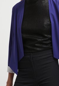 Lila, lätt kavaj med öppen front och trekvartsärmar, lager-på-lager över en blank svart figurnära topp. Strukturerade svarta byxor med hög midja.