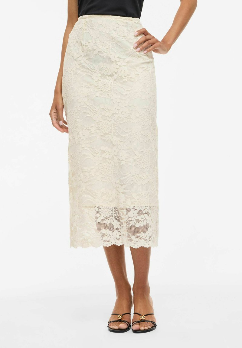 Jupe midi en dentelle crème avec motifs floraux, forme structurée, taille cintrée et ourlet festonné. Associée à des sandales noires à lanières.