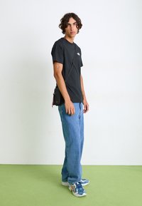 Sort t-shirt med et logo, blå denimjeans og blå sneakers. En lille sort crossbody-taske med en pink rem hænger fra skulderen.