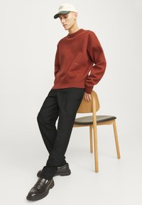 Pull en maille couleur rouille avec une coupe décontractée, associé à un pantalon noir et des mocassins noirs épais, le tout sur un fond simple.