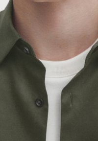 Camicia abbottonata verde oliva con colletto. Sotto, è visibile una t-shirt bianca a girocollo. Tessuto liscio in cotone.