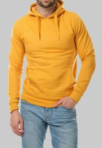 Hoodie jaune en coton, avec une poche frontale, un capuchon à cordon et des poignets côtelés. Porté avec un jean bleu clair.
