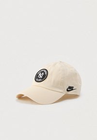 Beżowa czapka z daszkiem z czarną naszywką New York Yankees z przodu oraz czarnym logo Nike z boku na jednolitym jasnym tle.