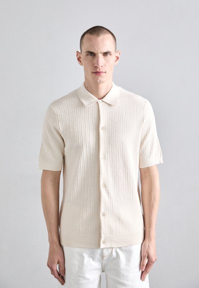 Chemise polo en tricot crème à manches courtes avec une texture côtelée, devant boutonné et col classique. Portée avec un pantalon blanc.